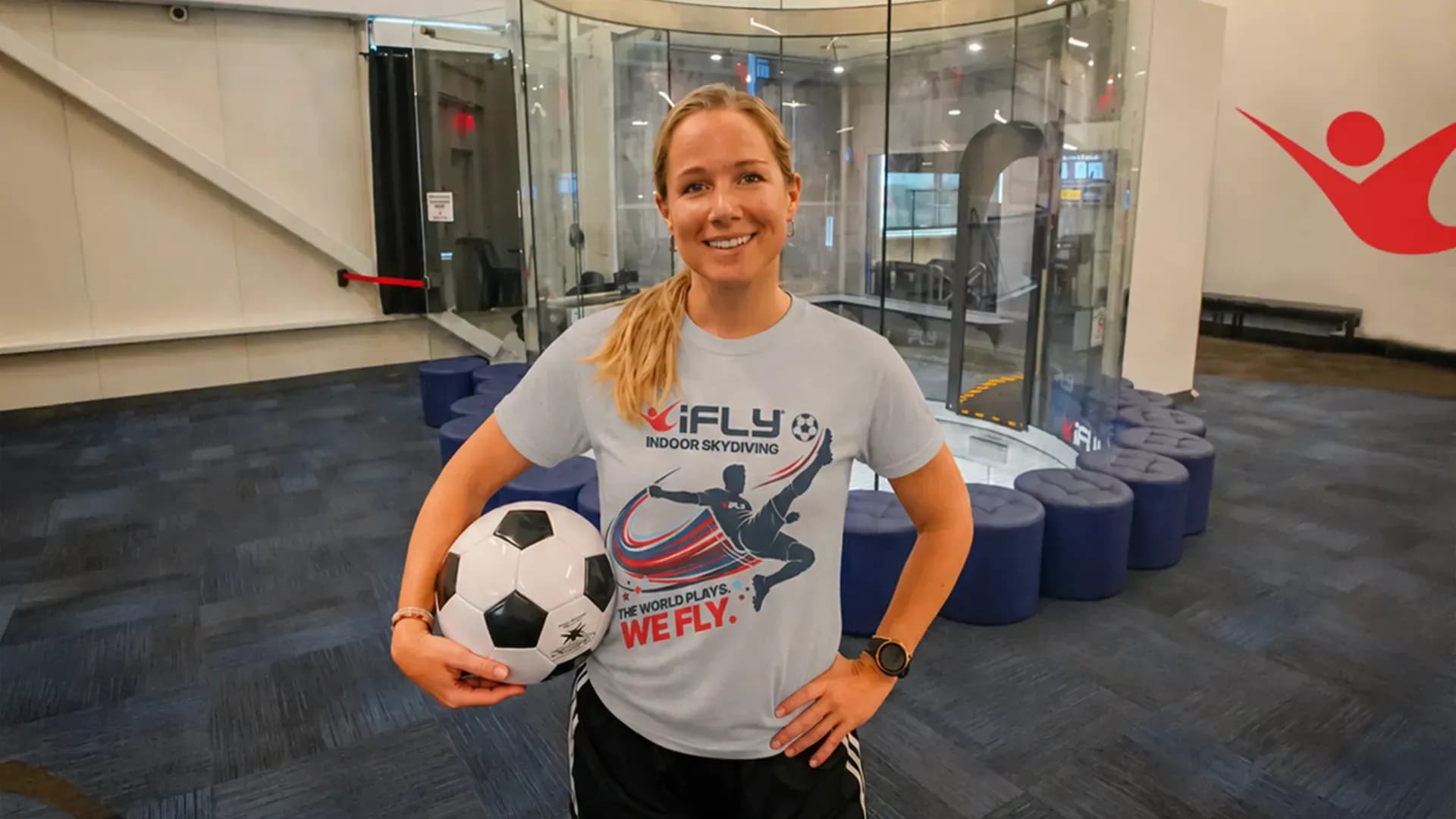 Smiling woman in an iFLY shirt holding a soccer ball inside a wind tunnel facility.