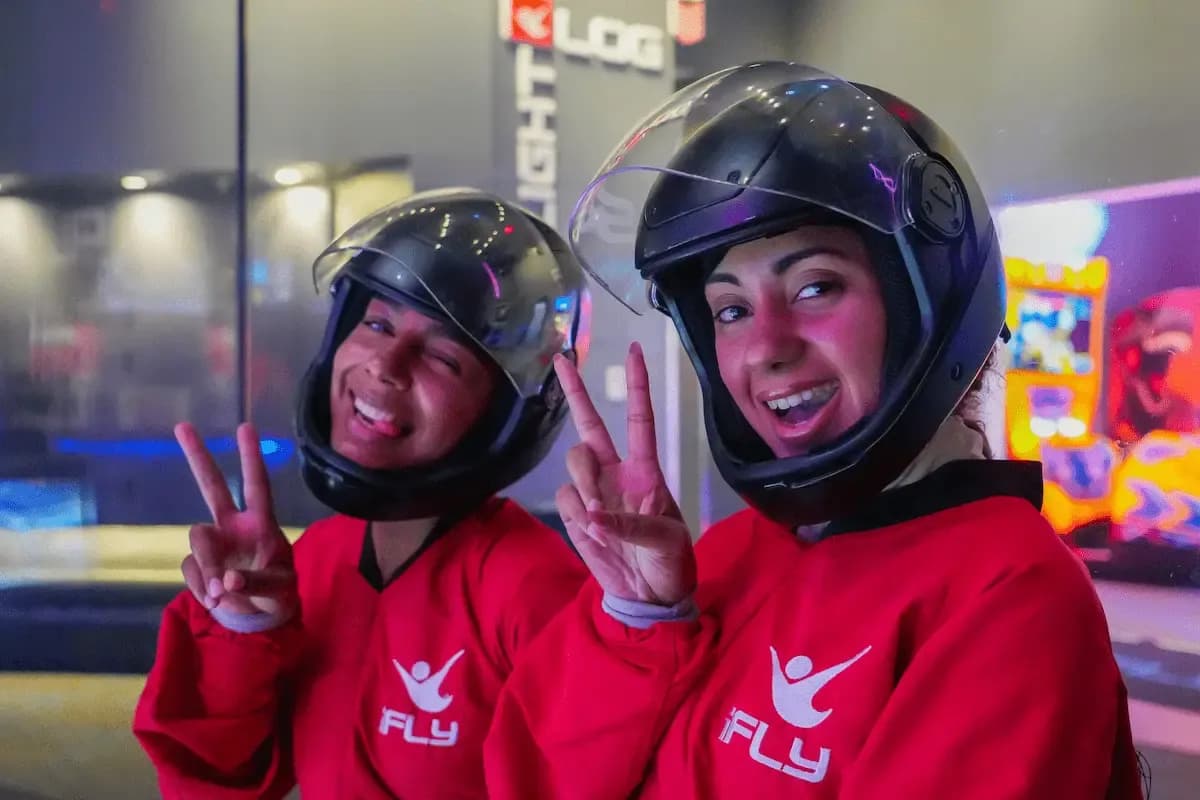 Two smiling women in red iFLY suits and helmets make peace signs indoors.