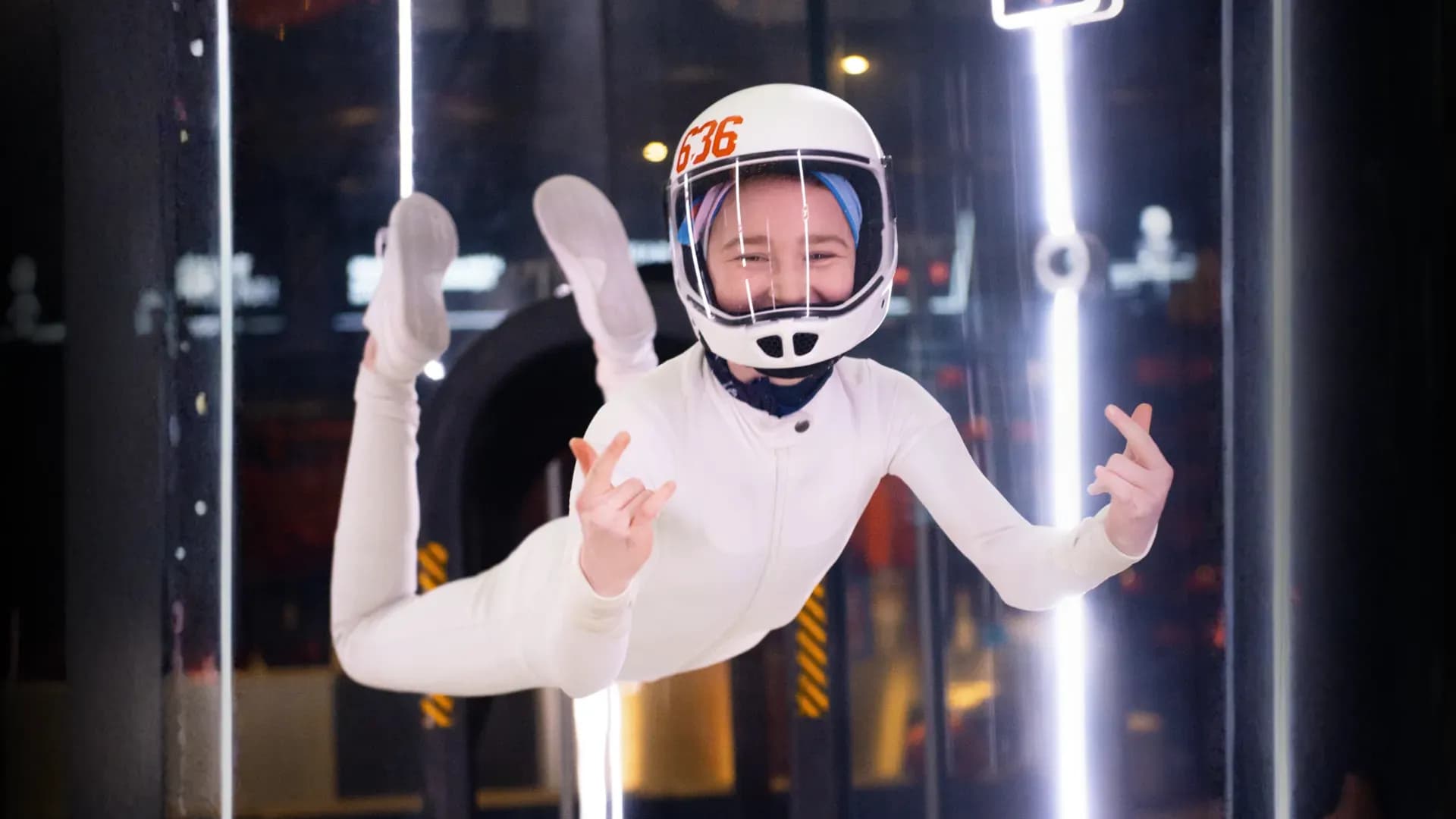 Smiling person in white suit and helmet floats in a wind tunnel, making heart gestures.