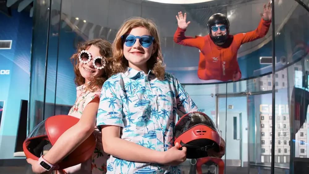 Smiling girls hold helmets while someone flies in an indoor skydiving wind tunnel.