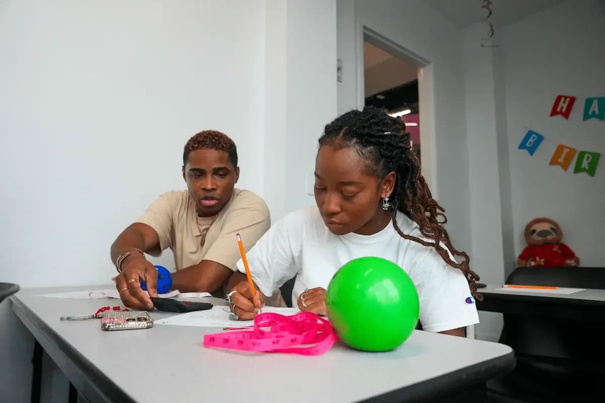 Two students in a classroom, one writing with a green ball and measuring tape.